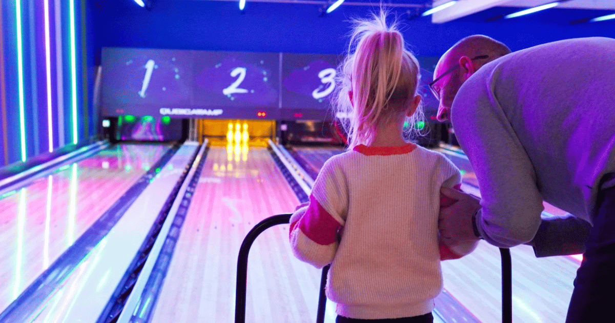 Man Teaching Young Girl To Roll A Ball Down Bowling Alley Lane (1)
